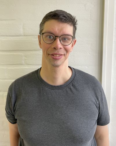 Aapolla on lyhyet ruskeat hiukset, silmälasit ja harmaa t-paita.